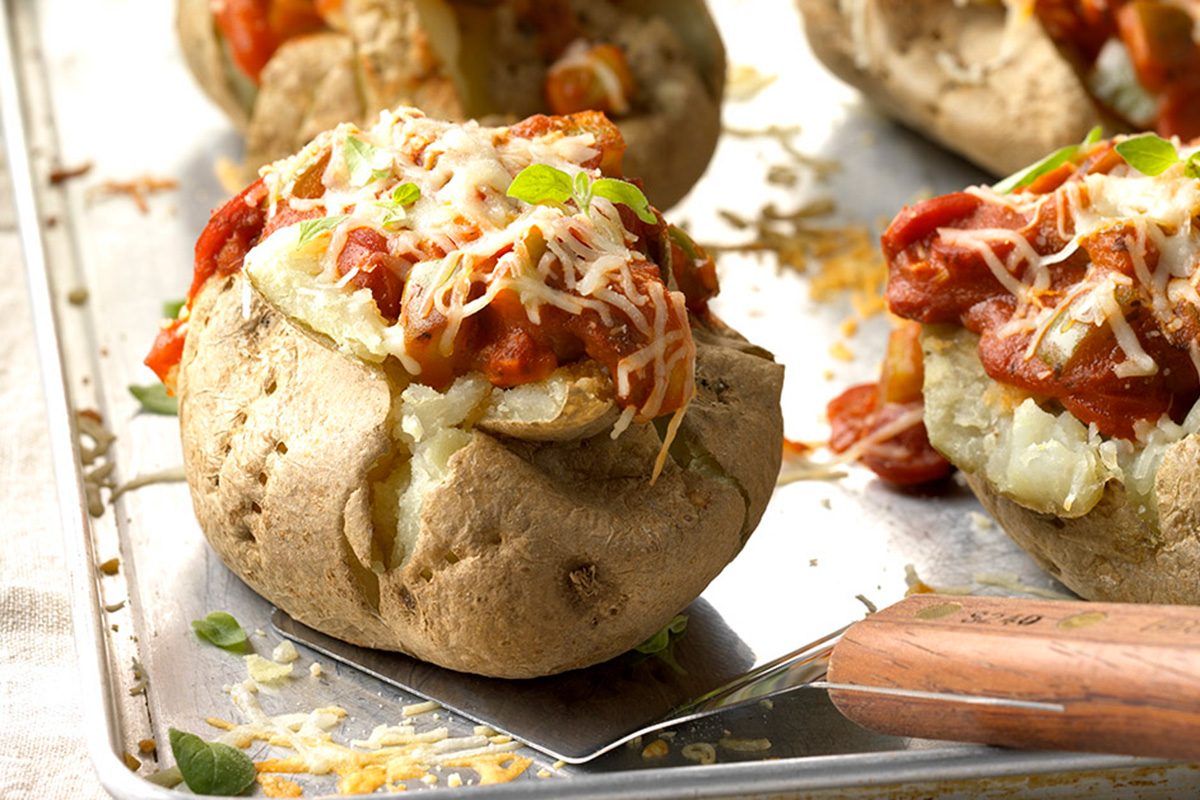 Baked potatoes split open and topped with tomato sauce, melted shredded cheese, and fresh herbs, served on a metal baking sheet with a serving spatula.