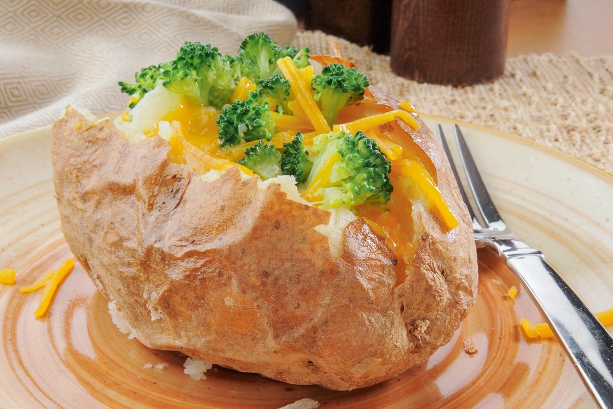 A baked potato topped with melted cheddar cheese and broccoli florets sits on a ceramic plate, with a fork and knife beside it.