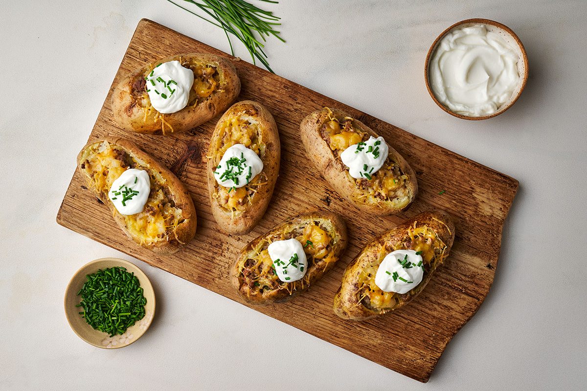 Six twice-baked potatoes topped with sour cream and chopped chives are arranged on a wooden serving board. A small bowl of chopped chives and a bowl of sour cream are nearby.