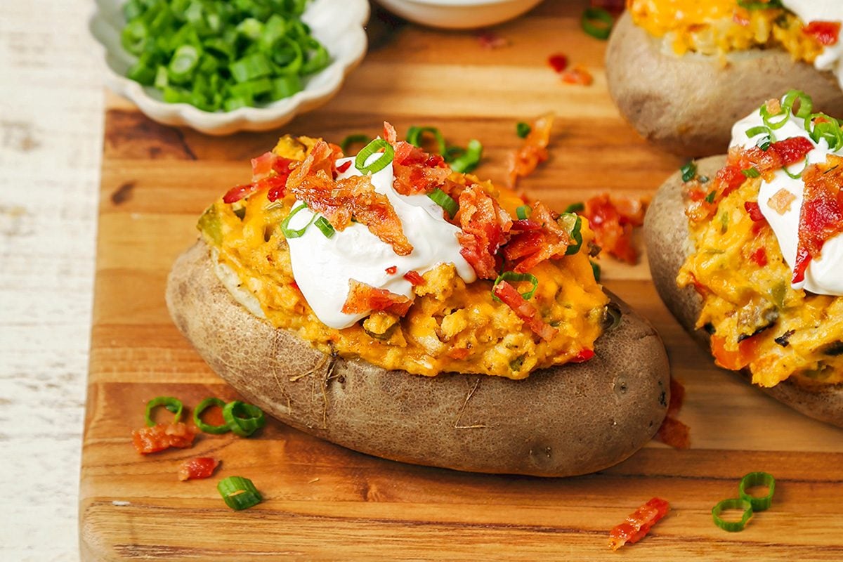 A baked potato topped with melted cheese, sour cream, chopped green onions, and crispy bits of bacon sits on a wooden board, with more potatoes and a bowl of green onions in the background.
