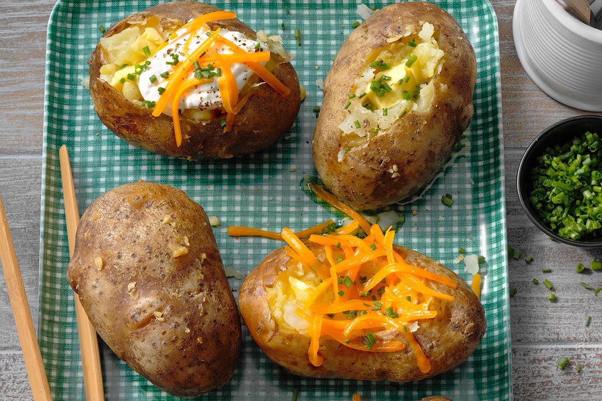 Four baked potatoes on a green checkered plate, two topped with shredded cheddar cheese, one with sour cream and chives, and one plain with chopped herbs. Chopsticks are placed beside the plate.