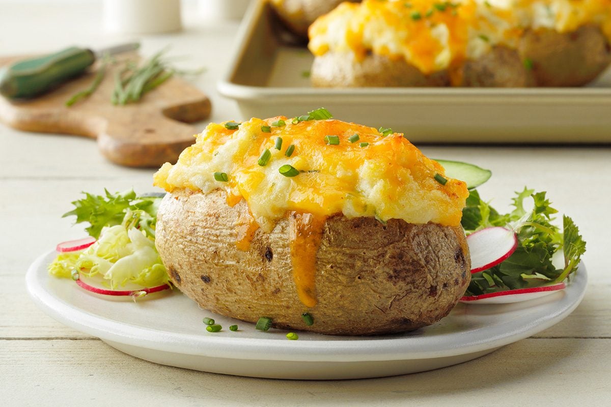 A baked potato topped with melted cheese and chopped chives is served on a white plate with a side of fresh greens, radish slices, and cucumber.