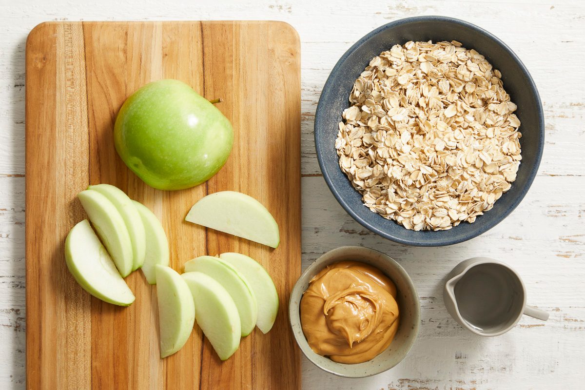 Overhead photo of ingredients on a wooden cutting board including sliced green apple, oats, peanut butter, and water in a small bowl.