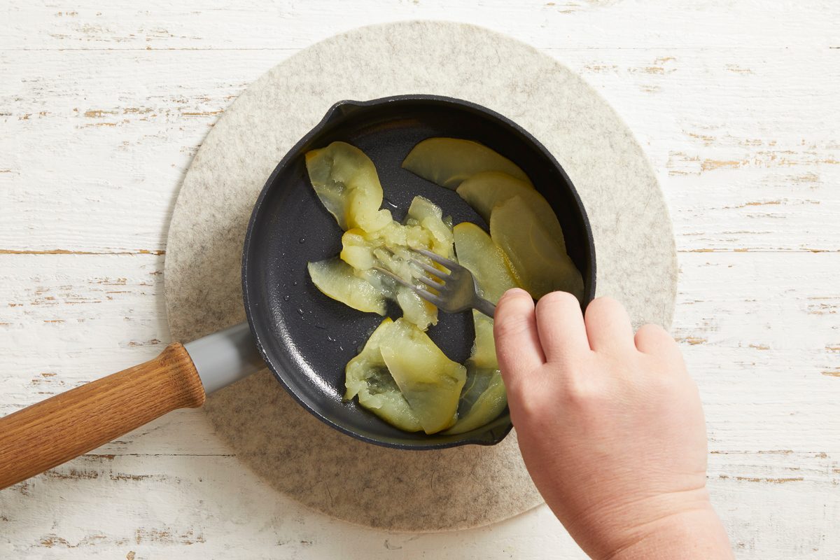 Overhead image of apple slices being sautéed in a pan on a stovetop, stirred by hand with a wooden spatula.