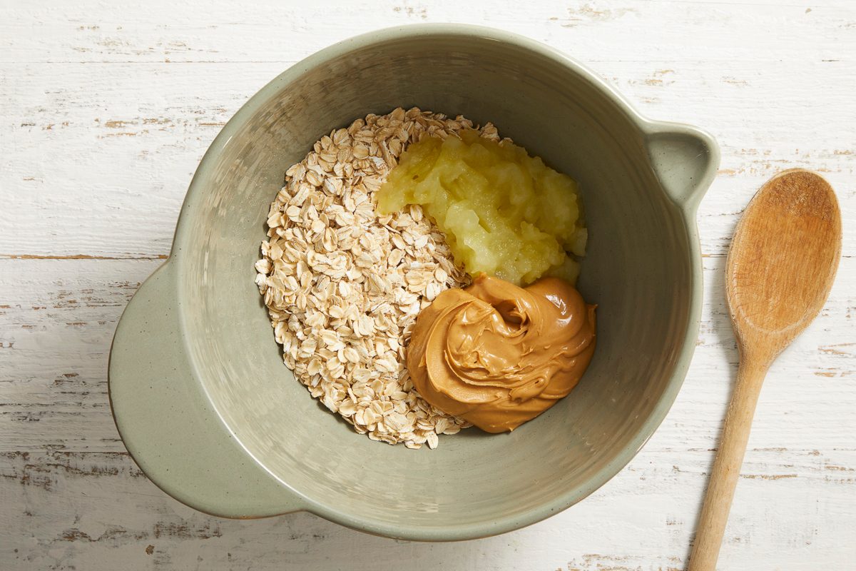 Overhead view of a mixing bowl with oats, sautéed apple, and peanut butter ready to be stirred together with a wooden spoon beside.