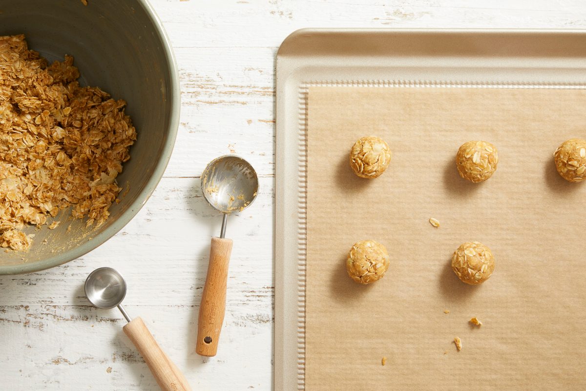 Overhead image of scooped dough portions on a parchment-lined baking sheet, shaped into balls using cookie scoops nearby.