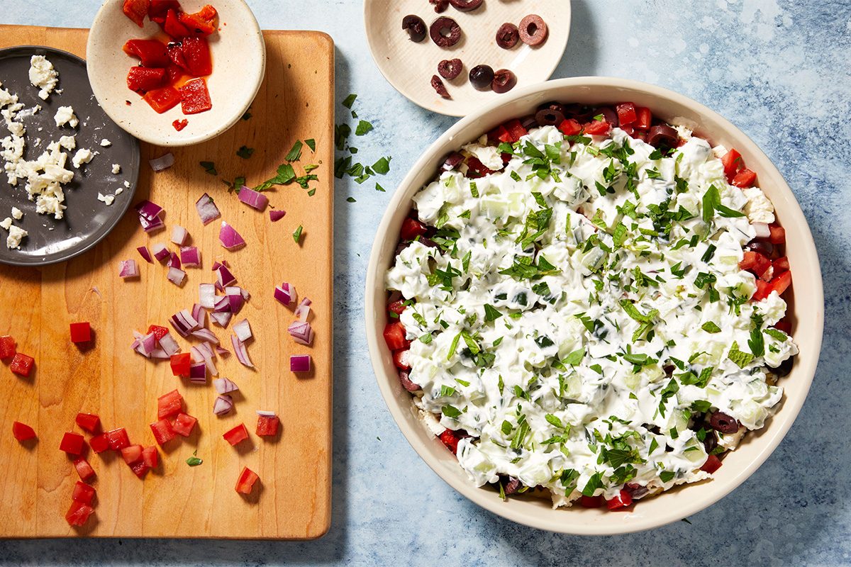 Overhead shot of Greek yogurt and cucumber tzatziki-style mixture being added on top of the layered hummus dip in a bowl, with scattered toppings around.