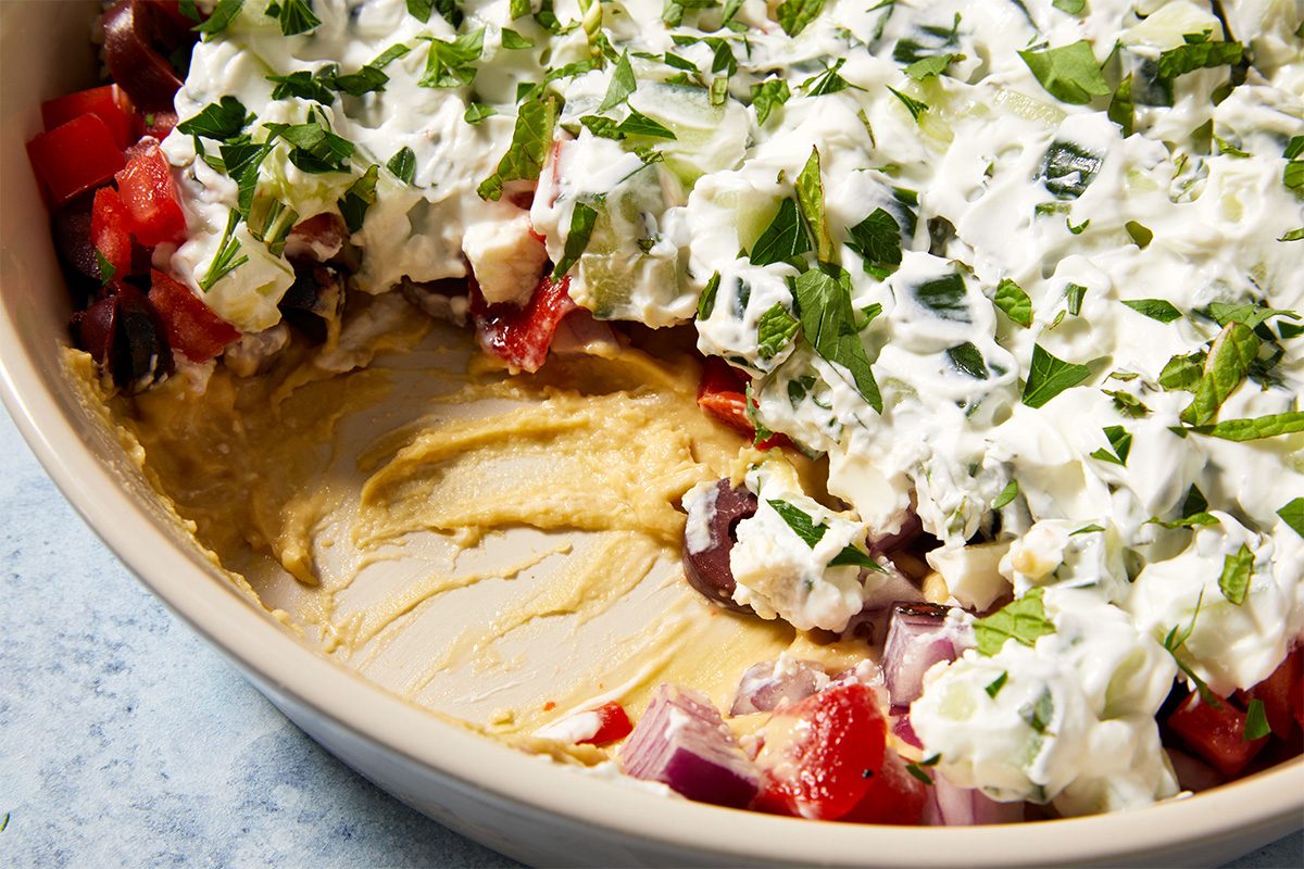 Close up shot of partially eaten Mediterranean layered dip, revealing the different colorful layers