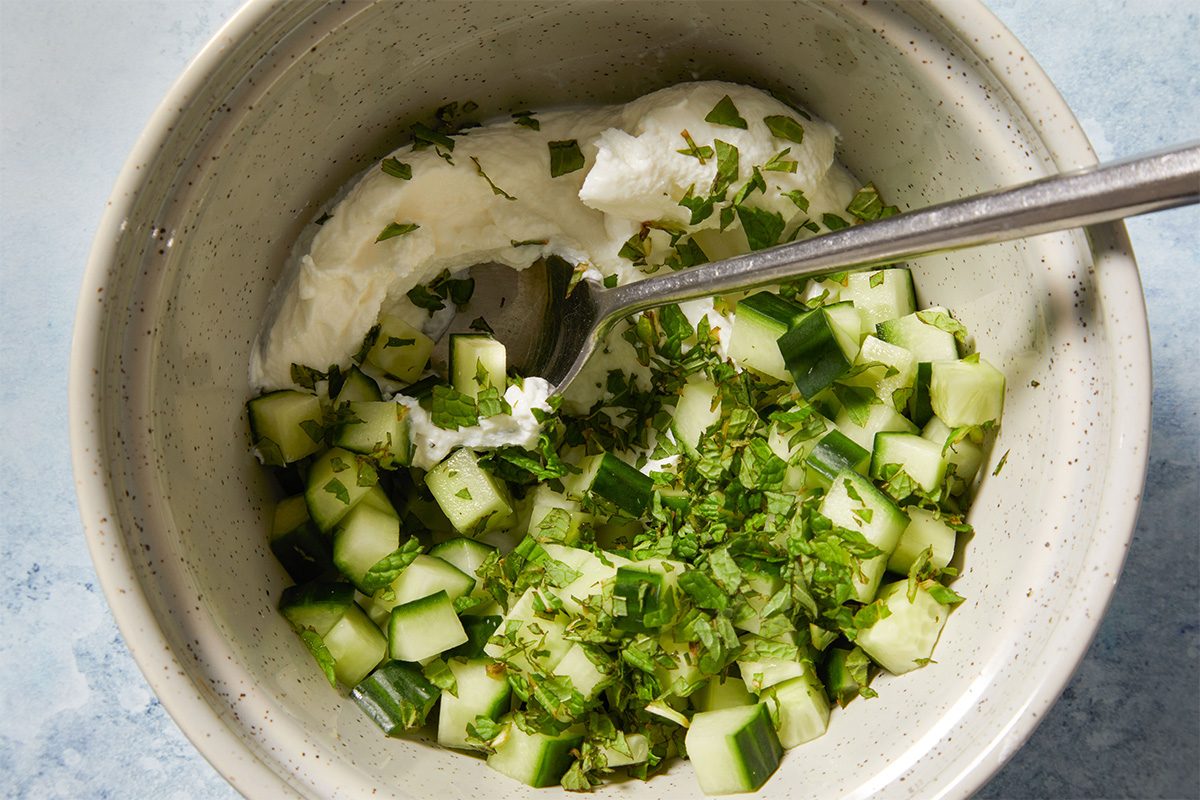 Overhead shot of diced cucumber and herbs being mixed into strained yogurt in a metal bowl with a spoon inside.