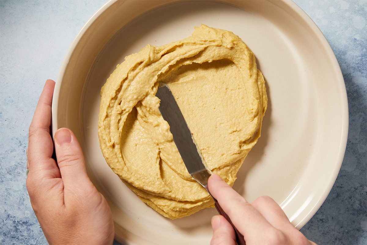 Overhead close-up shot of hands spreading a thick layer of hummus into the base of a shallow beige bowl.