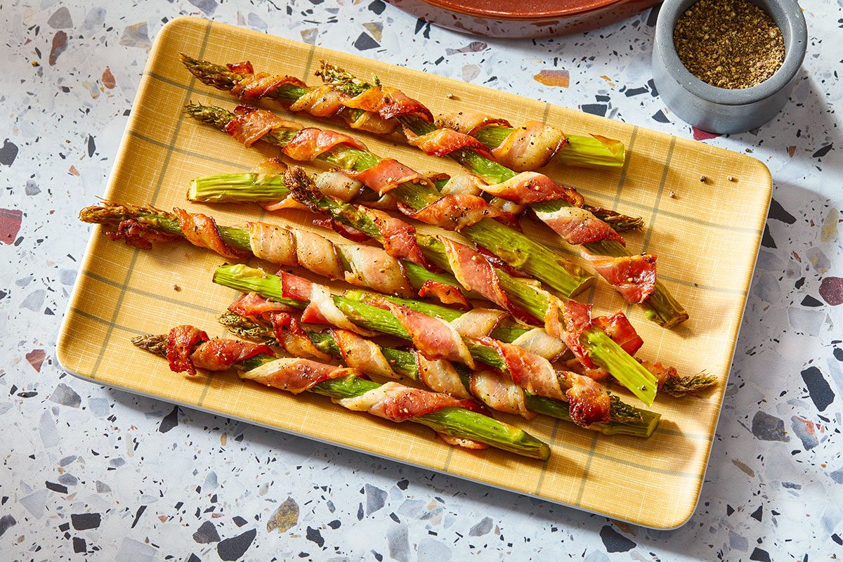 A horizontal overhead view shows bacon-wrapped asparagus arranged on a rectangular platter with seasoning visible.