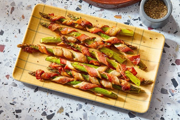 A horizontal overhead view shows bacon-wrapped asparagus arranged on a rectangular platter with seasoning visible.