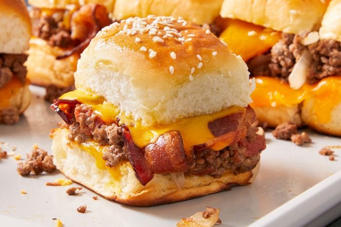 Bacon Cheeseburger Sliders on a white tray with one in focus in center with melted cheese dripping out of it.