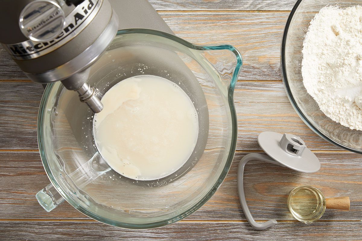Overhead shot of a stand mixer with a glass bowl containing milk and yeast on a wooden surface, with a bowl of flour