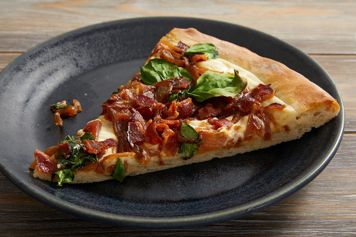 Closeup shot of a slice of bacon and spinach pizza served on a dark plate
