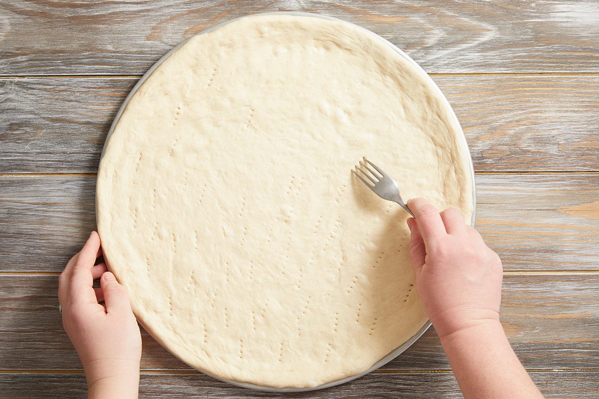 Overhead shot of hands using a fork to poke holes in raw pizza dough