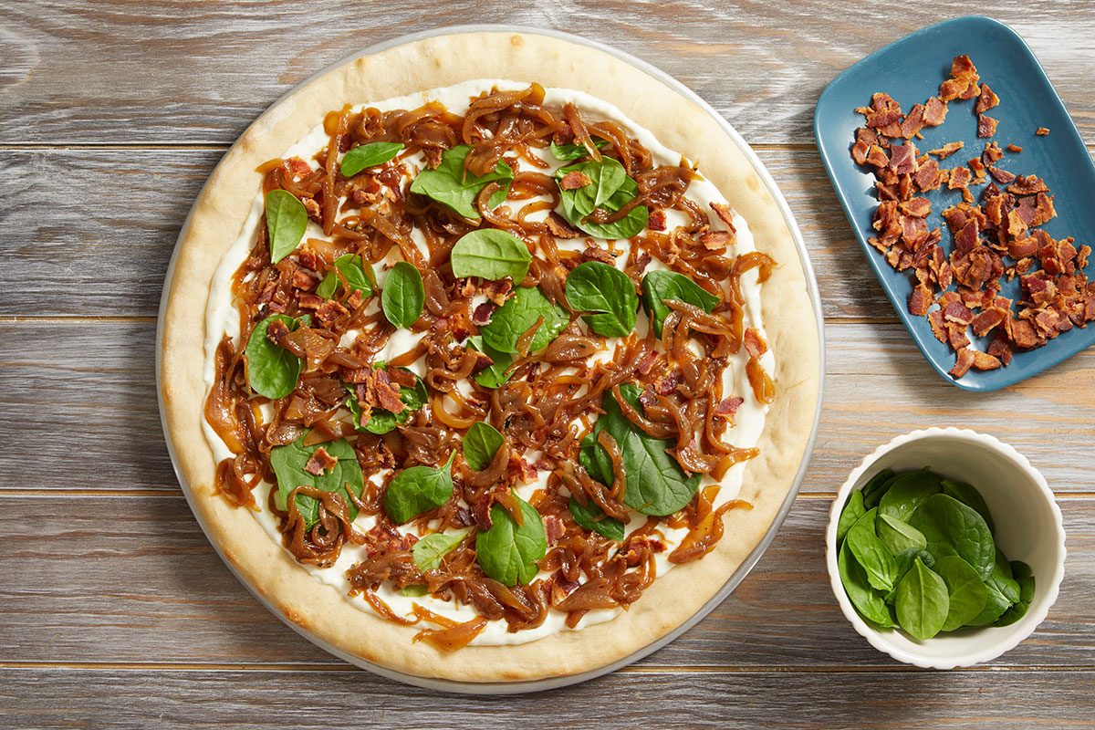 Overhead shot of a pizza topped with caramelized onions, crispy bacon bits, and fresh spinach leaves