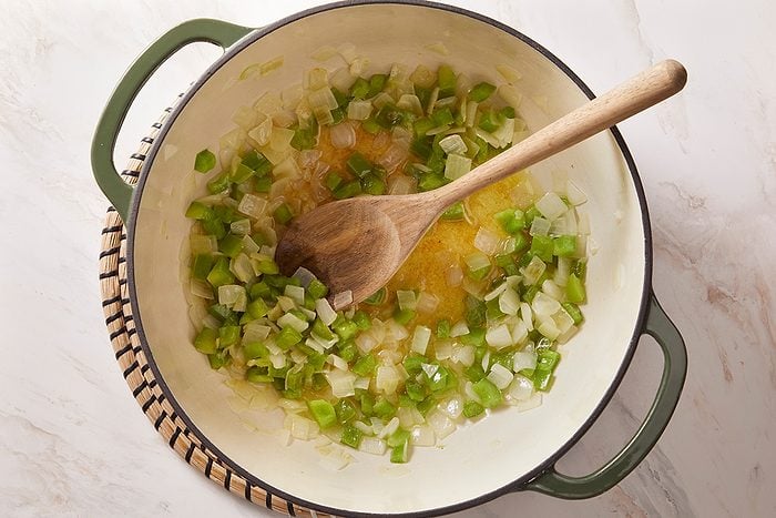A green Dutch oven on a white surface contains diced onions and green bell peppers being sautéed, stirred with a wooden spoon.