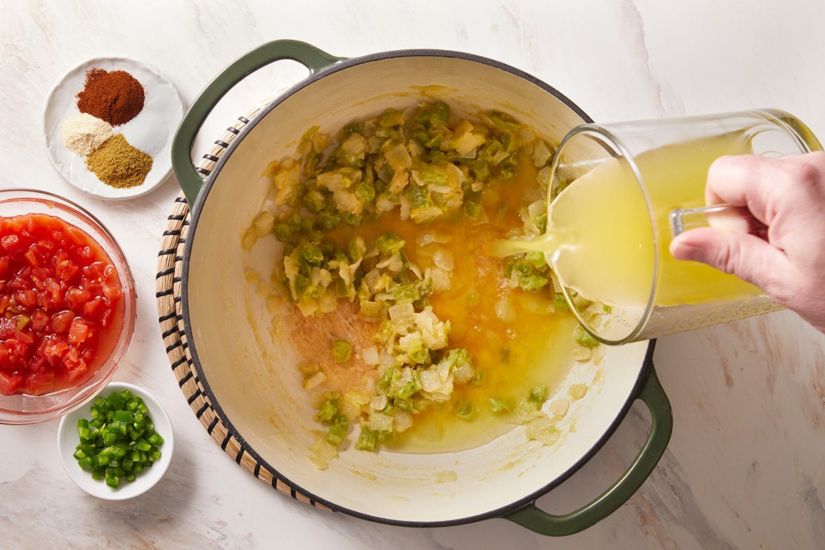 A hand pours broth into a pot with sautéed onions and peppers. Nearby are bowls containing chopped tomatoes, diced green peppers, and assorted spices on a white countertop.