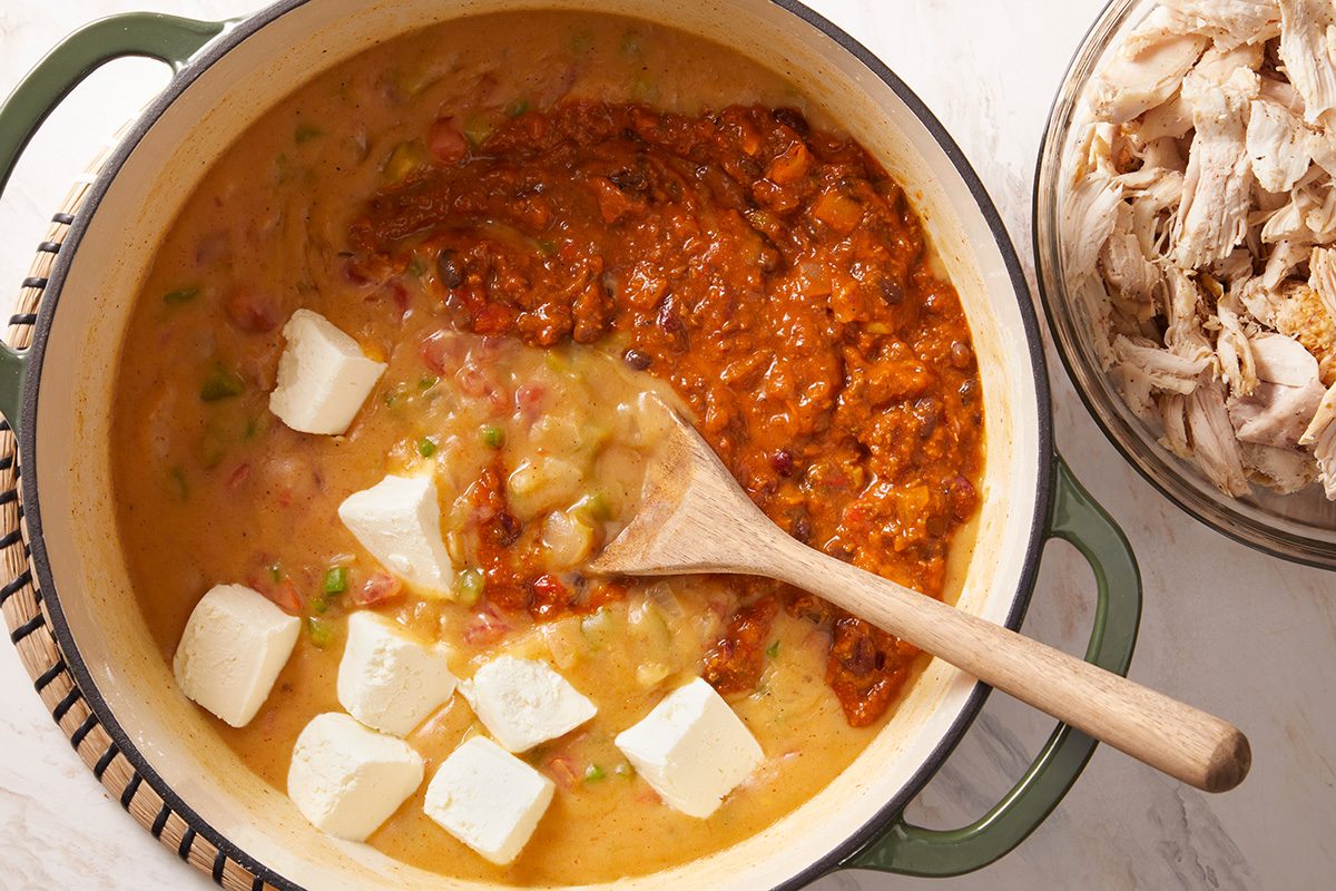 A pot filled with a creamy mixture, dollops of white cream cheese, and a red sauce being stirred with a wooden spoon; a bowl of shredded chicken sits nearby.