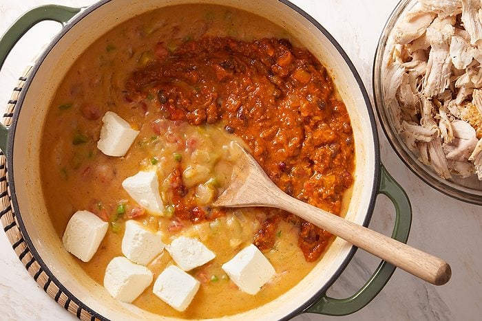 A pot filled with a creamy mixture, dollops of white cream cheese, and a red sauce being stirred with a wooden spoon; a bowl of shredded chicken sits nearby.