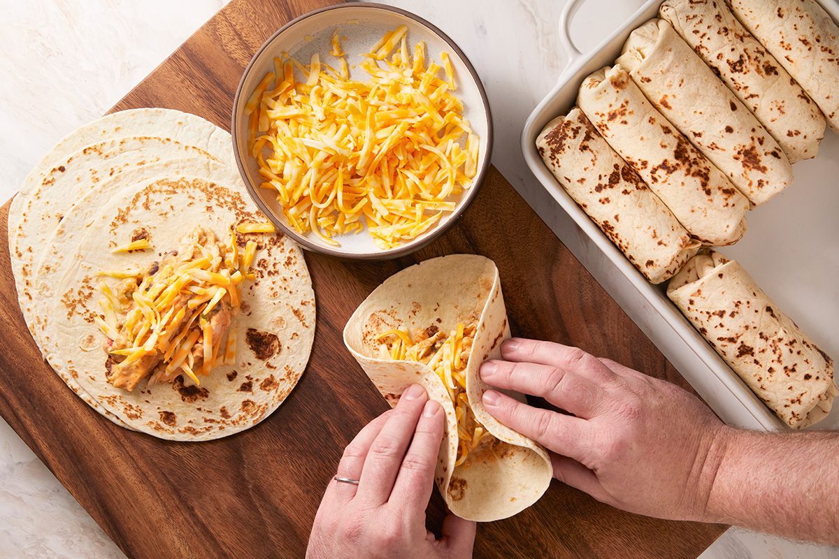 Hands fill a tortilla with shredded cheese on a wooden board. Nearby are more tortillas, a bowl of shredded cheese, and a baking dish with rolled, filled tortillas.