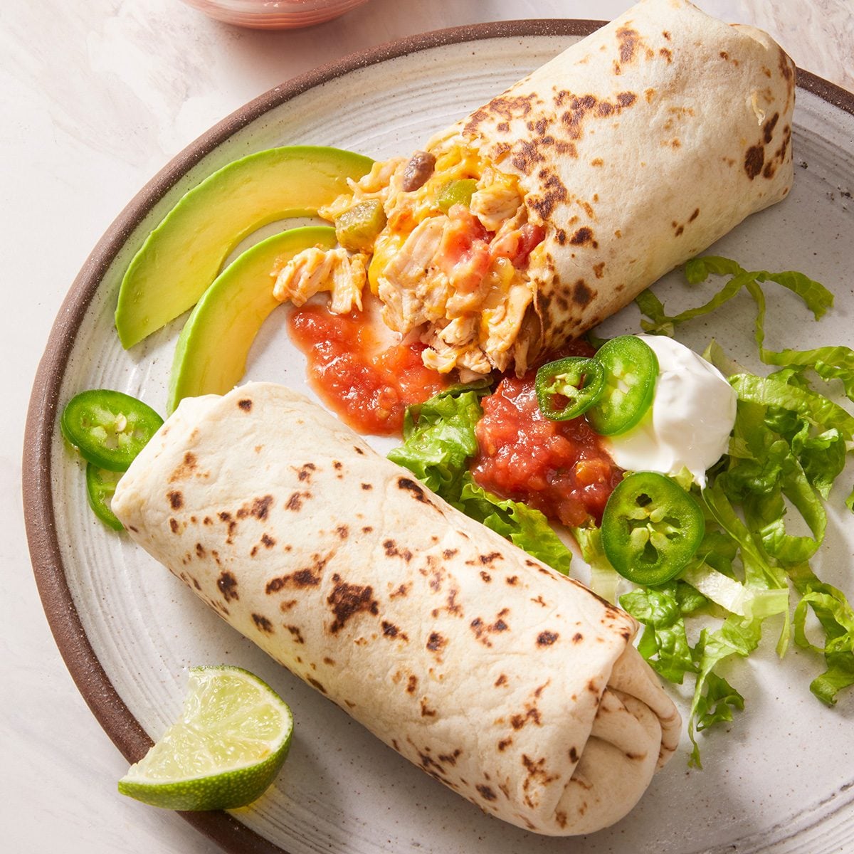 A plate with two breakfast burritos, one cut open showing eggs and salsa, served with sliced avocado, jalapeños, lettuce, sour cream, and a lime wedge.