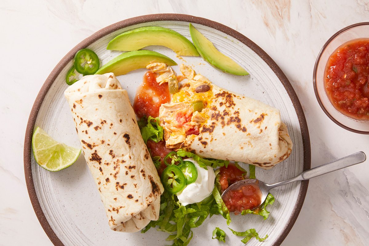 A plate with two burritos, sliced avocado, lime wedge, jalapeño slices, shredded lettuce, sour cream, and salsa, with a bowl of salsa and a spoon on the side.