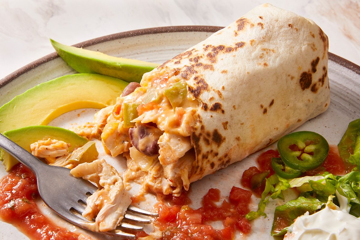 A chicken burrito with beans and cheese is partially cut open on a plate, served with slices of avocado, jalapeños, lettuce, and salsa. A fork is holding a bite of the burrito.