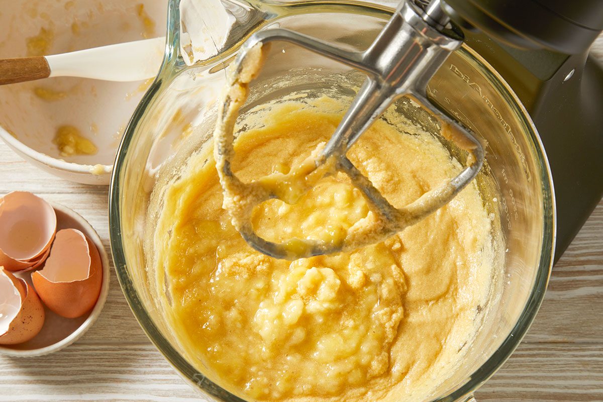 A horizontal overhead image shows a glass mixing bowl with a handheld mixer blending a pale yellow batter, with cracked eggs and baking ingredients arranged nearby.