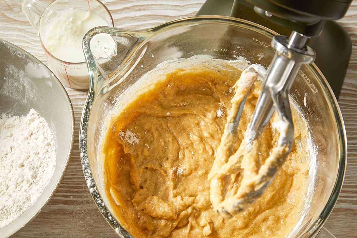 A horizontal overhead image shows thick banana batter being mixed in a glass bowl with a hand mixer, with flour and additional ingredients nearby.