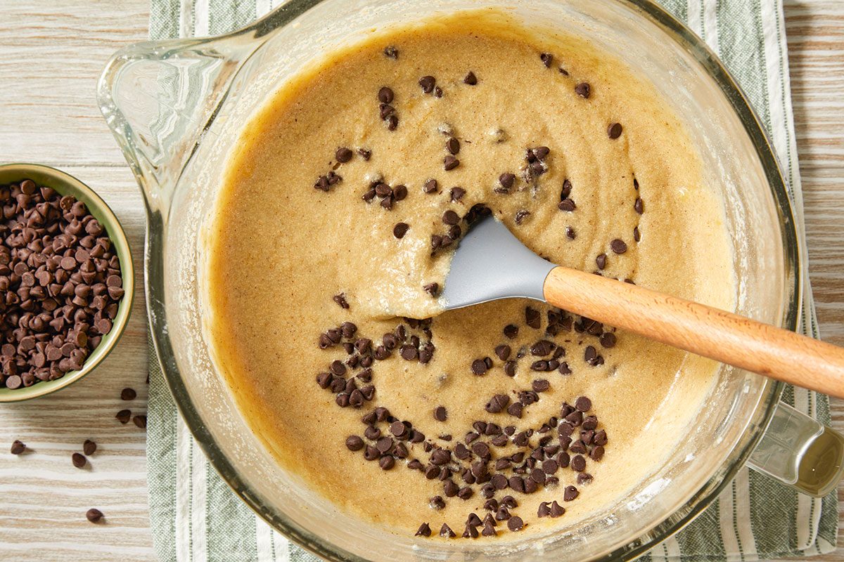 A horizontal overhead image shows chocolate chips scattered into banana batter in a glass bowl, with a spatula resting in the mixture.