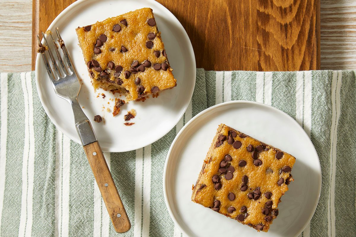 A horizontal overhead image shows individual squares of banana chocolate chip cake plated on small dishes with forks on a striped cloth.