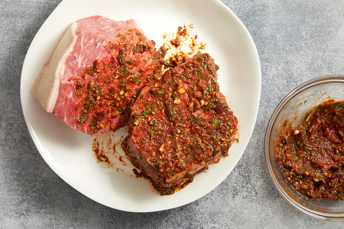 A raw steak coated with a thick herb and spice marinade sits on a white plate, with a small bowl of extra marinade nearby on a gray surface.