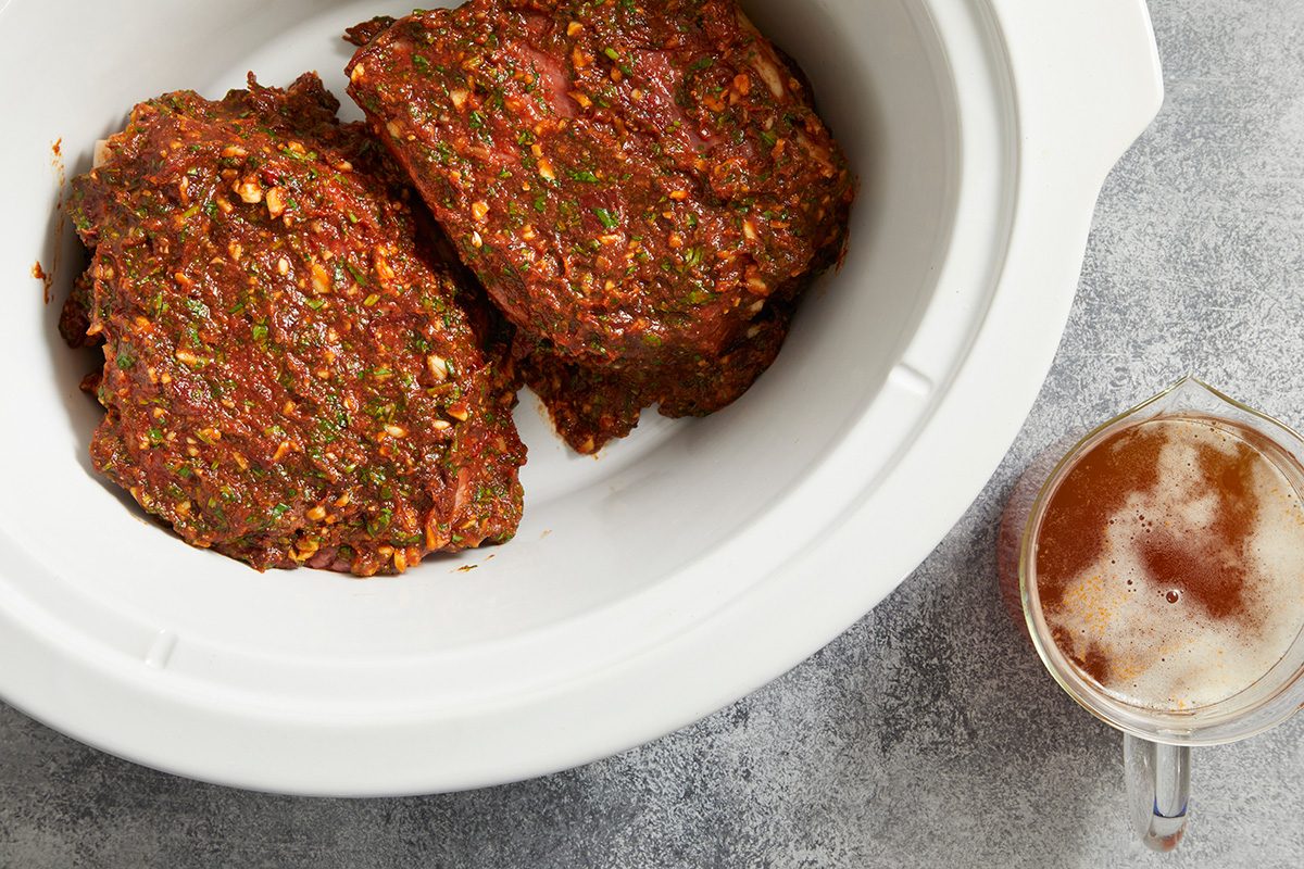 Two seasoned pieces of meat sit in a white slow cooker, with a measuring cup of frothy liquid placed beside it on a gray countertop.