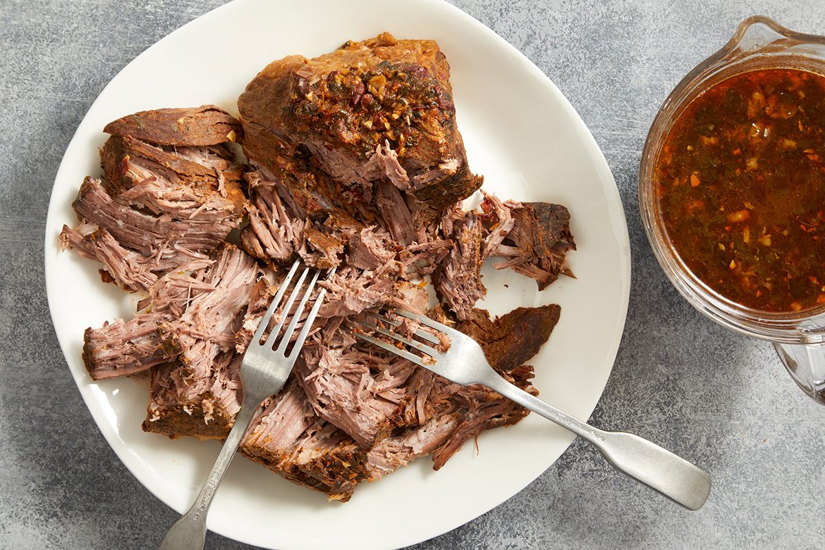 A plate of shredded beef with two forks, next to a glass container of brown sauce on a gray surface.