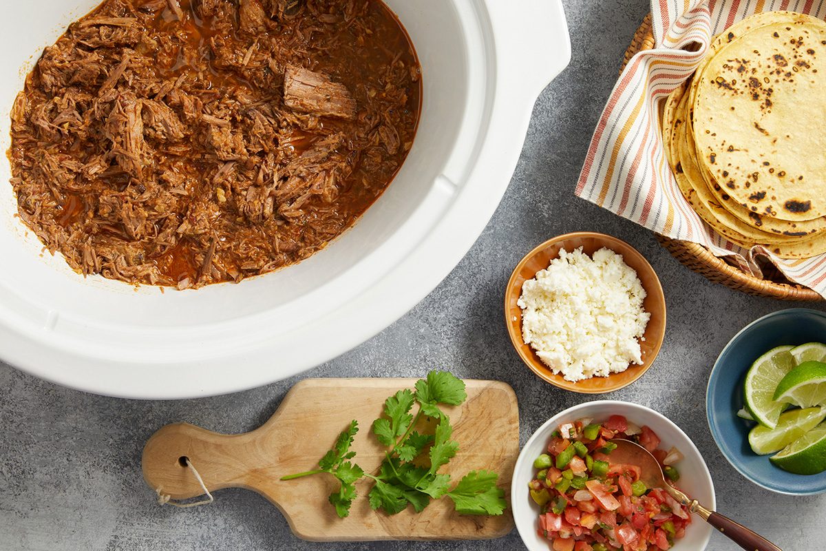 Shredded meat in a slow cooker, with bowls of crumbled cheese, salsa, lime wedges, tortillas, and a cutting board with fresh cilantro on a gray surface.