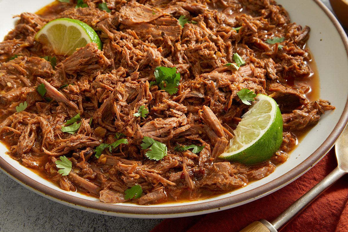 A bowl filled with shredded barbacoa beef in a rich sauce, garnished with fresh cilantro and lime wedges.