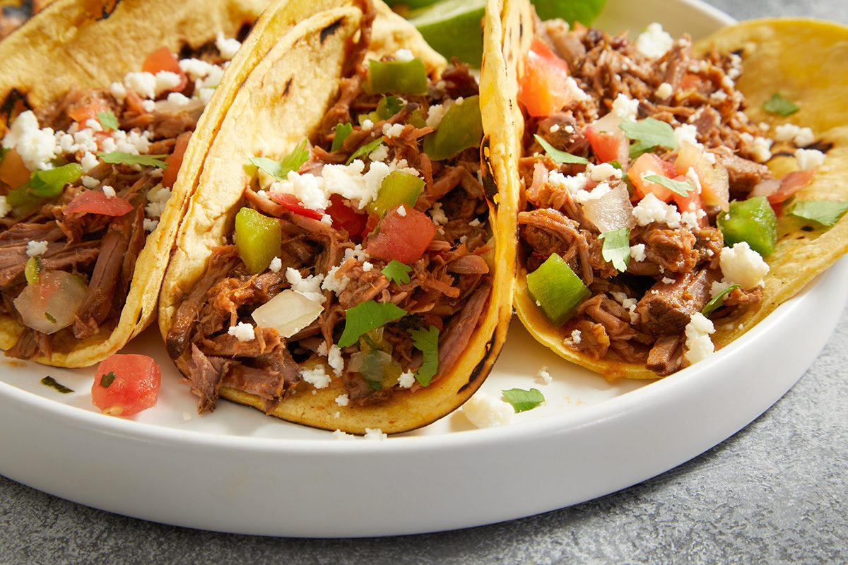 Three corn tortillas filled with shredded beef, diced tomatoes, green bell peppers, onions, and crumbled cheese are arranged on a white plate. Lime wedges are partly visible in the background.