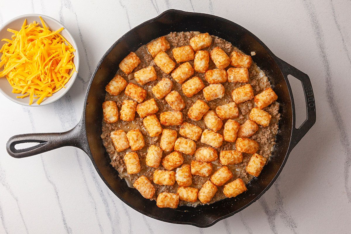 Frozen tater tots are arranged in an even layer over the beef mixture in the skillet.
