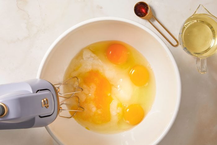 A horizontal overhead image shows eggs and sugar being mixed in a large bowl with a hand mixer during muffin batter preparation.
