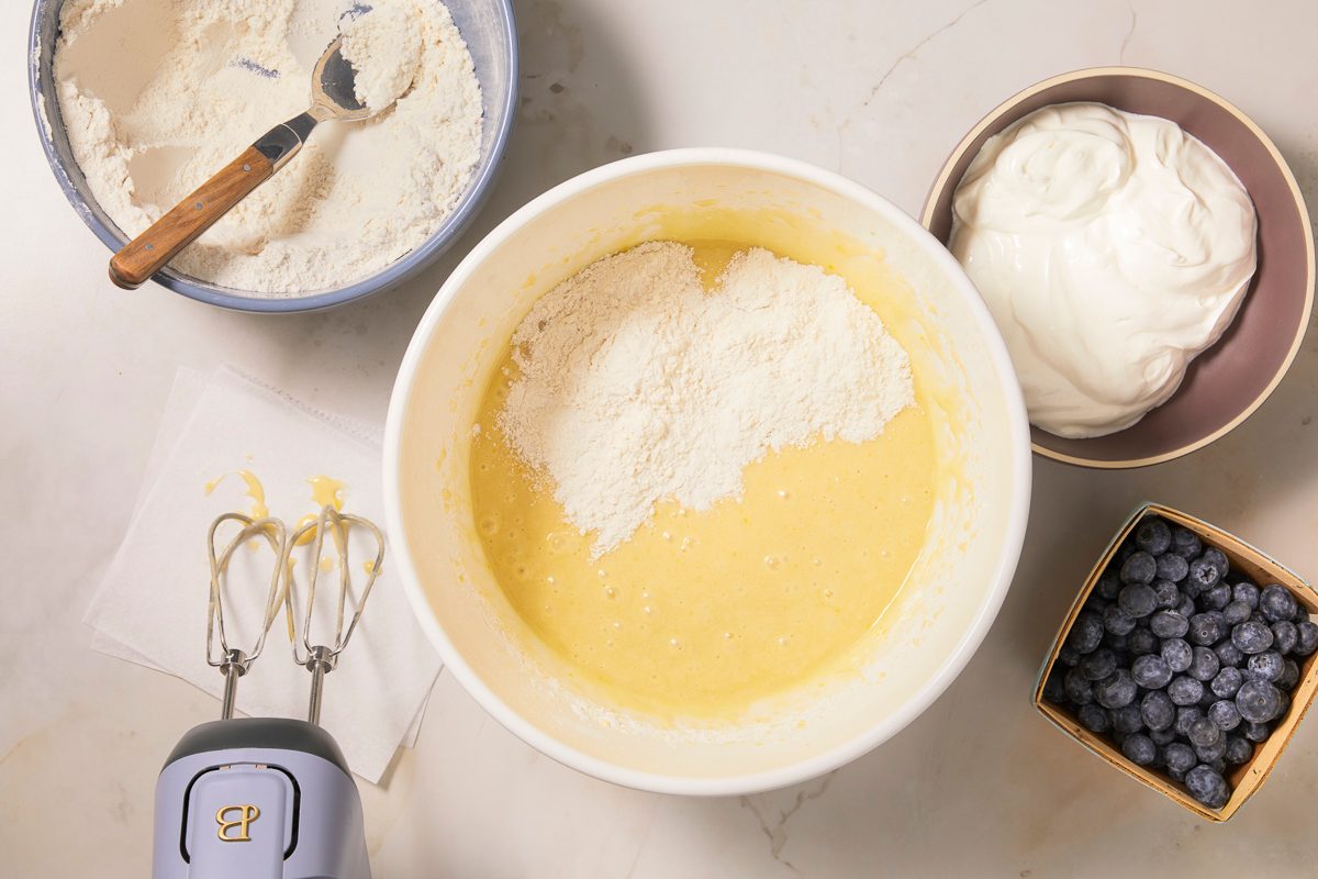 A horizontal overhead image shows dry ingredients being added to a wet mixture alongside bowls of flour, sour cream, and blueberries during batter preparation.