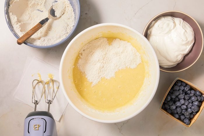 A horizontal overhead image shows dry ingredients being added to a wet mixture alongside bowls of flour, sour cream, and blueberries during batter preparation.
