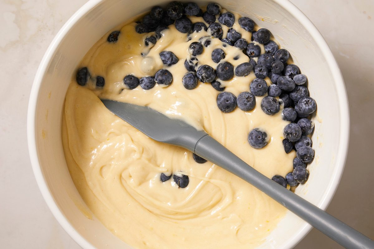 A horizontal close-up image shows fresh blueberries being folded into thick muffin batter with a spatula.