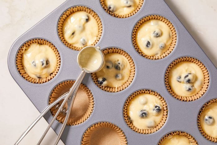 A horizontal overhead image shows muffin batter portioned into lined muffin cups in a baking tin using a scoop.