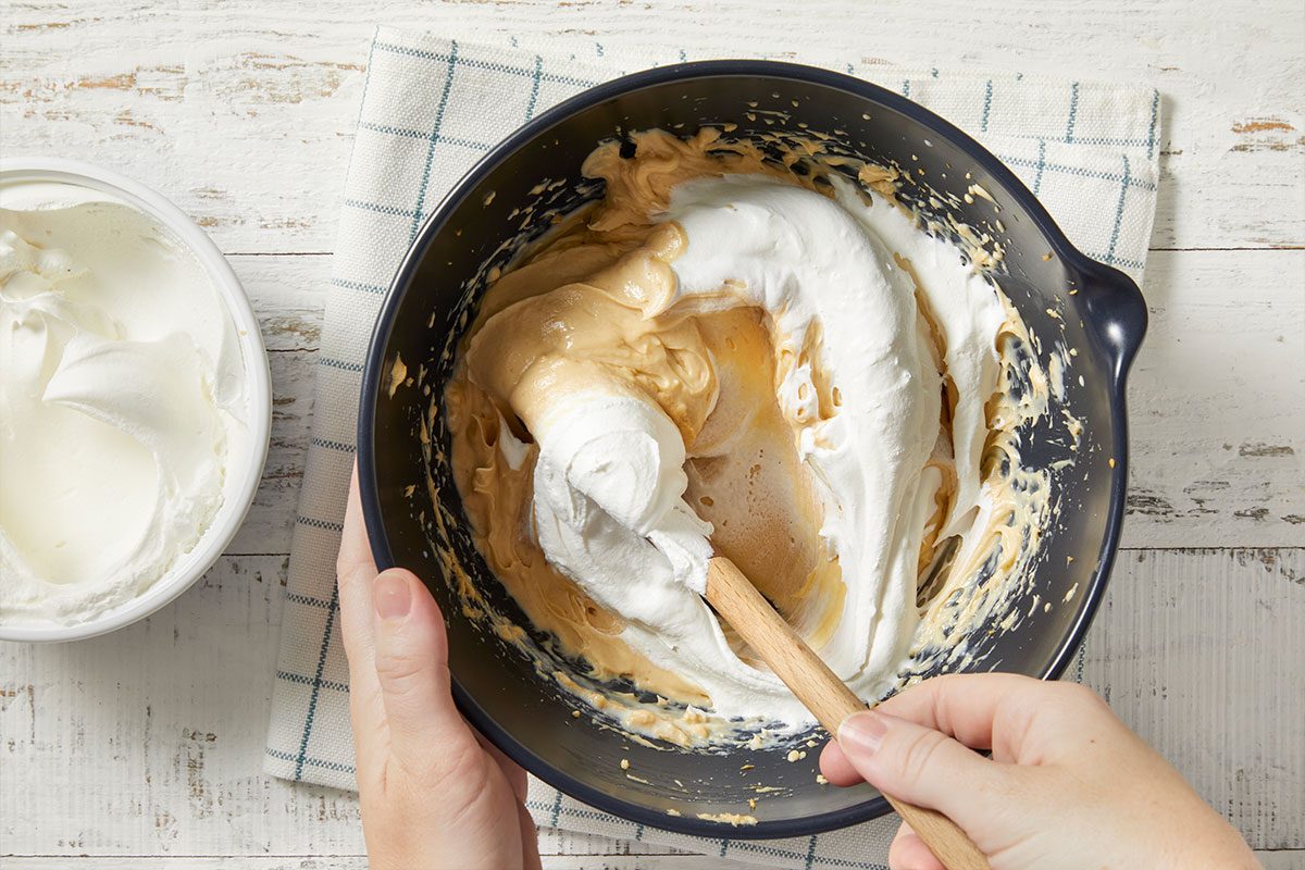 Overhead horizontal image of a wooden spoon mixing a light brown batter with fluffy white topping in a dark blue bowl being held. A bowl of the fluffy white topping sits on a white wooden surface alongside a blue and white gingham towel.