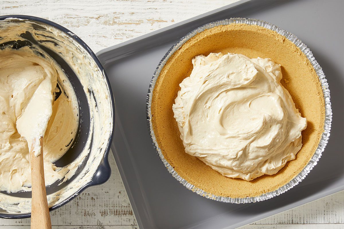 Overhead horizontal image of pie filling being poured into a graham cracker crust. The filling is in a bowl next to the pie, and a wooden spatula is resting inside the bowl.