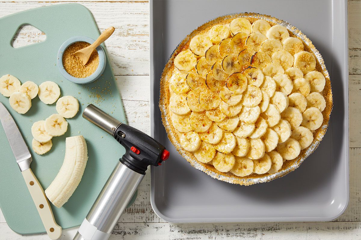 Overhead horizontal image of a banana pie with a kitchen torch and cutting board on a white wooden background. The banana slices are topped with sugar being torched, and next to the pie, bananas are being sliced on a teal cutting board with brown sugar in a small bowl.