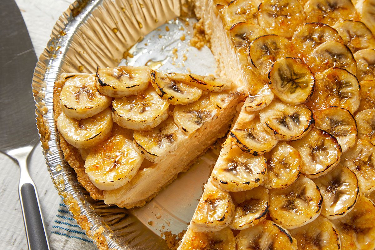 Overhead horizontal image of a sliced banana pie in a silver aluminum pie tin. The pie filling is visible, topped with caramelized banana slices. A metal serving utensil is on the side.
