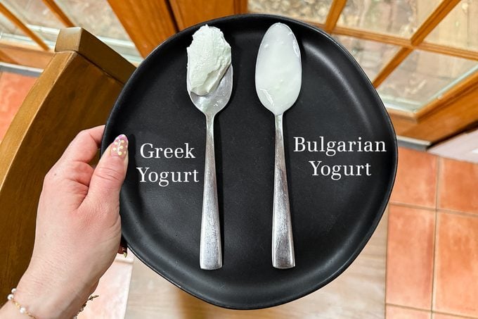 A hand holds a black plate with two spoons labeled "Greek Yogurt" and "Bulgarian Yogurt," each holding a scoop of yogurt. The Greek yogurt appears thicker than the Bulgarian yogurt.